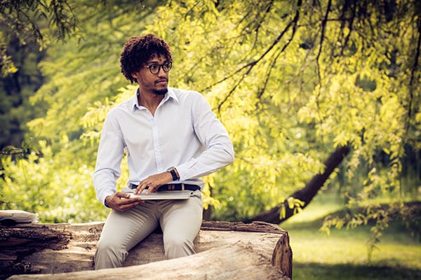 A man in formal attire with spectacles and a laptop sits sideways on a fallen tree trunk.